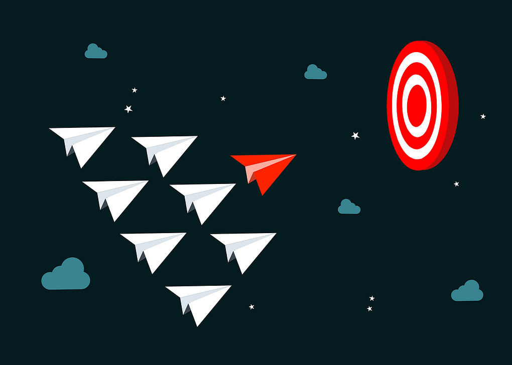 paper airplanes led by a red paper airplane. They are flying towards a dartboard. The dartboard signifies the goal and the red paper airplane exemplifies great leadership skills.