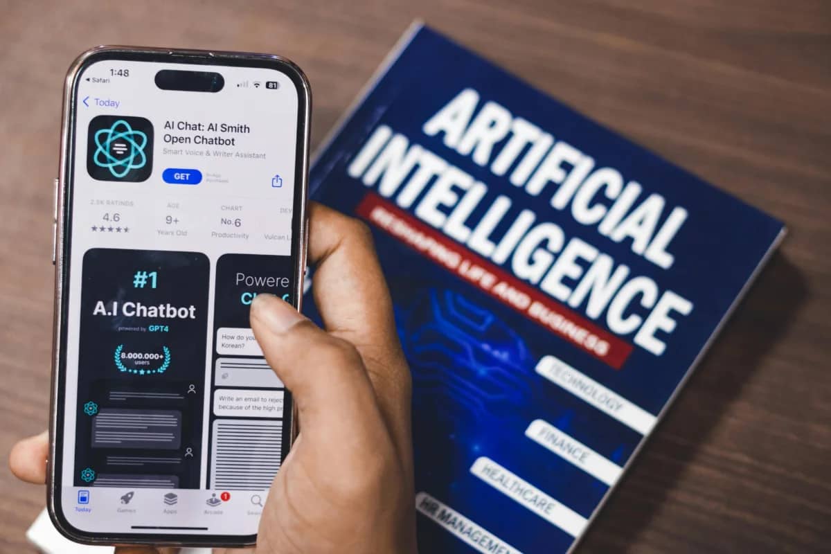 a person holding a phone showing an AI chatbot. In the background, is a book on artificial intelligence